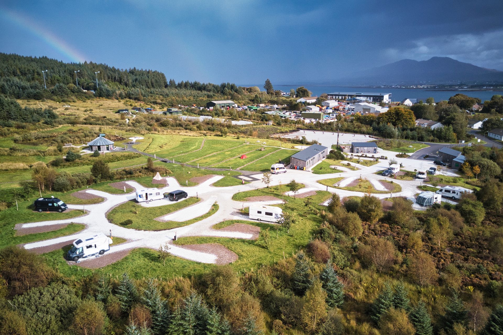 Aerial view of a coastal campsite with campervans parked on curving gravel pitches amid green lawns, small cabins and a central pavilion, bordered by dense woodland; a faint rainbow arches over the trees and distant mountains rise across the bay.