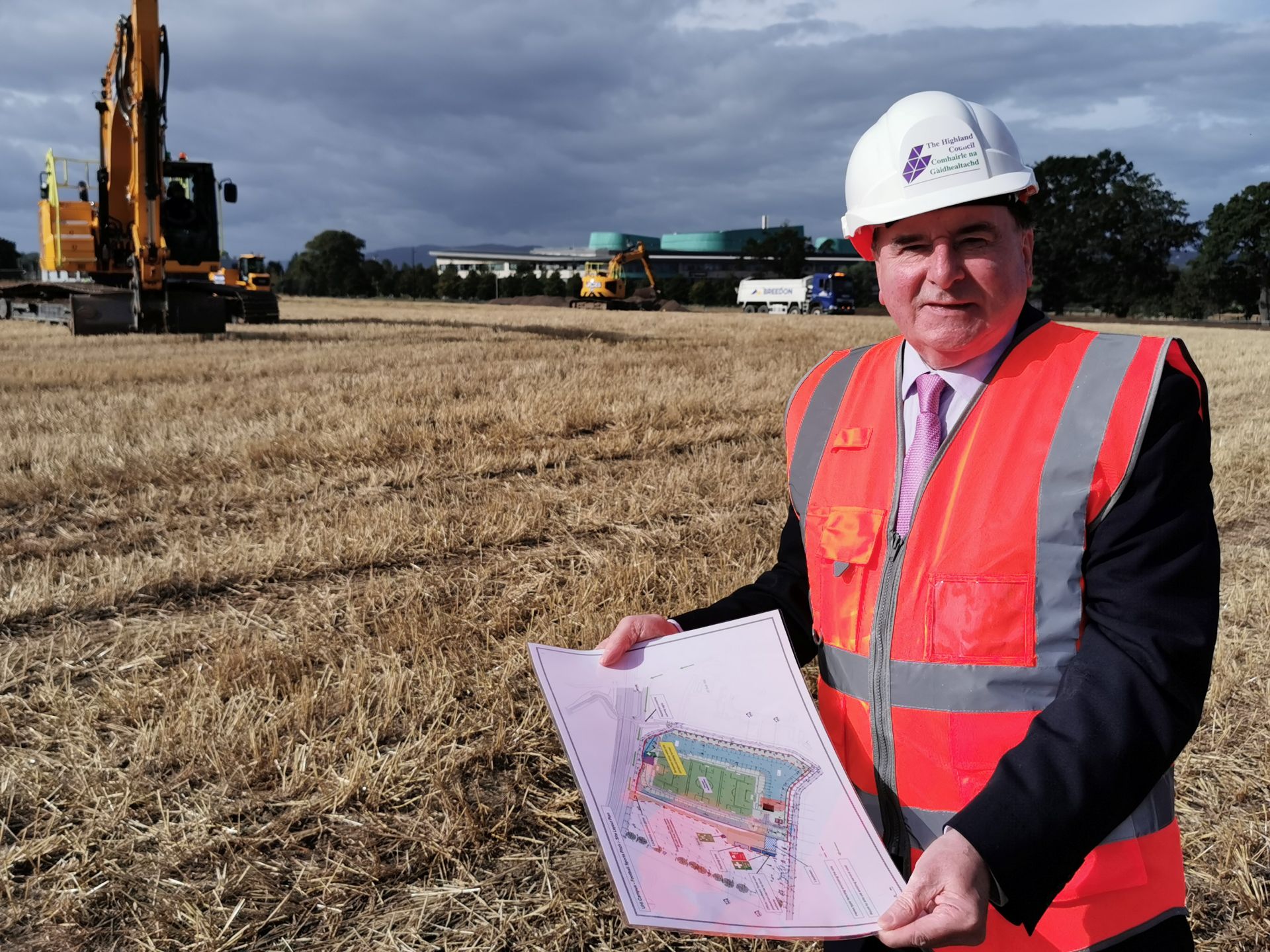 Man in a white hard hat labeled "The Highland Council" and an orange high-visibility vest holding site plans in a stubbled field with excavators and a truck preparing a construction site under a cloudy sky.
