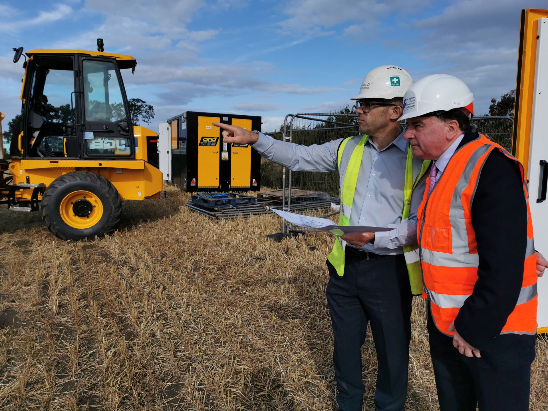 Two men wearing hard hats and high-visibility vests review plans while one points toward construction equipment and portable cabins during an on-site visit in a harvested field.
