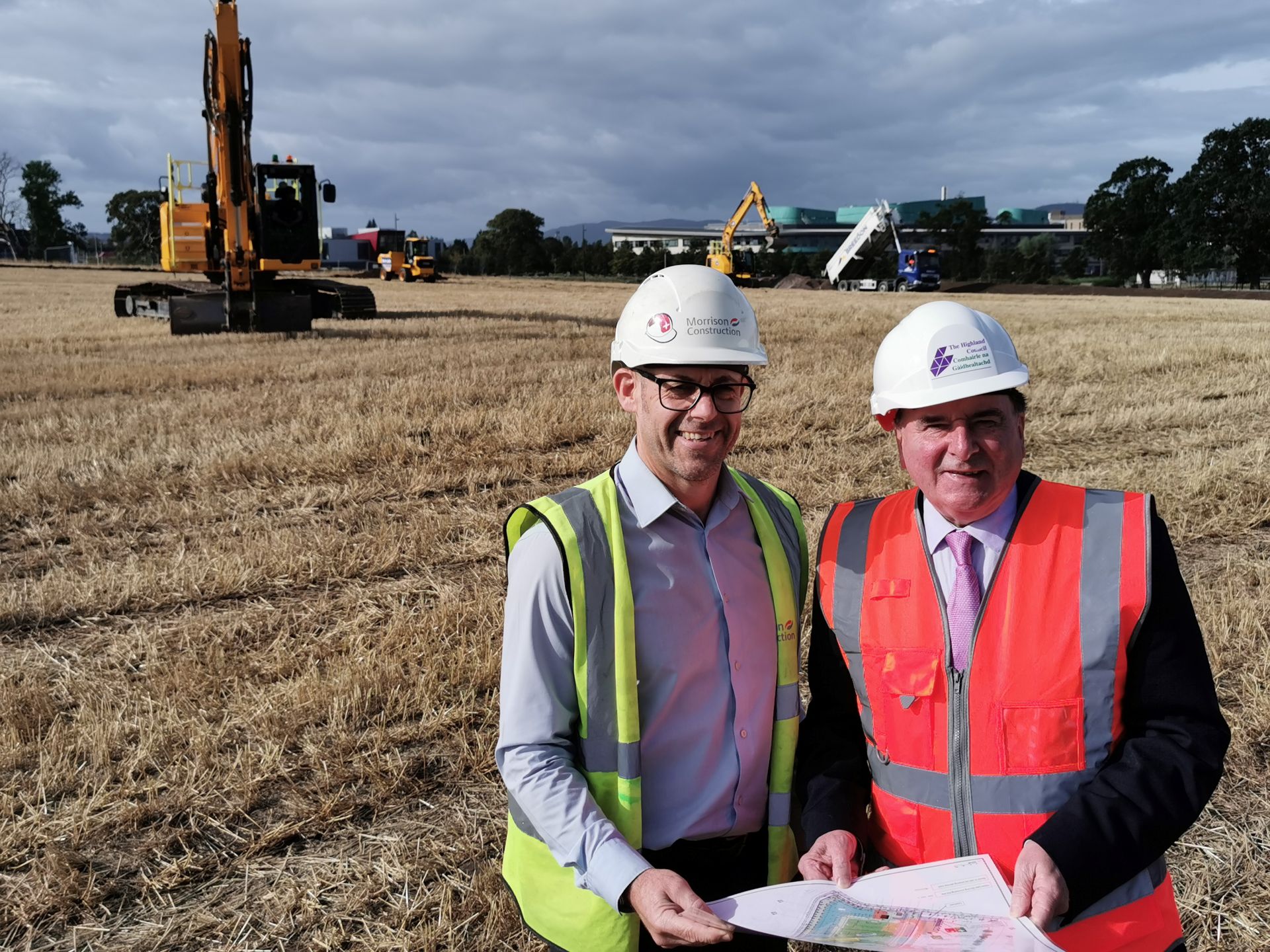 Two men in hard hats and high-visibility vests review site plans in a harvested field while excavators and a dump truck work in the background under a cloudy sky.
