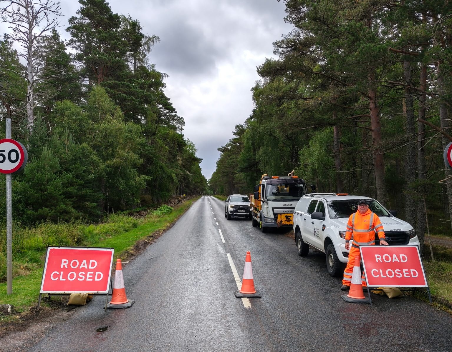 A road bordered by trees. On the road is road closed signs, along with cones, and a Council roads officer/vehicle.