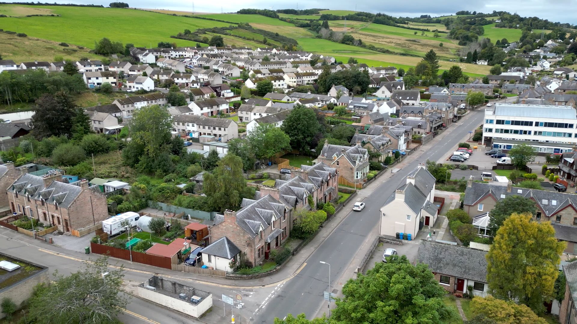 Aerial view of Dingwall surrounded by rolling green countryside. A main road runs through rows of houses, with residential streets, gardens, mature trees, and a large office building visible on the right.