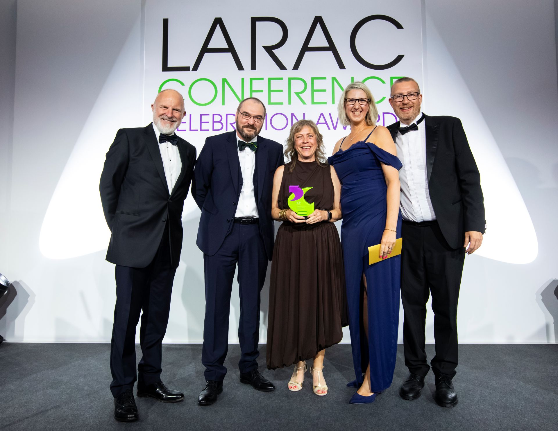 Five people in formalwear pose on stage at the LARAC Conference Celebration Awards, smiling as the woman in the centre holds a green-and-purple trophy.