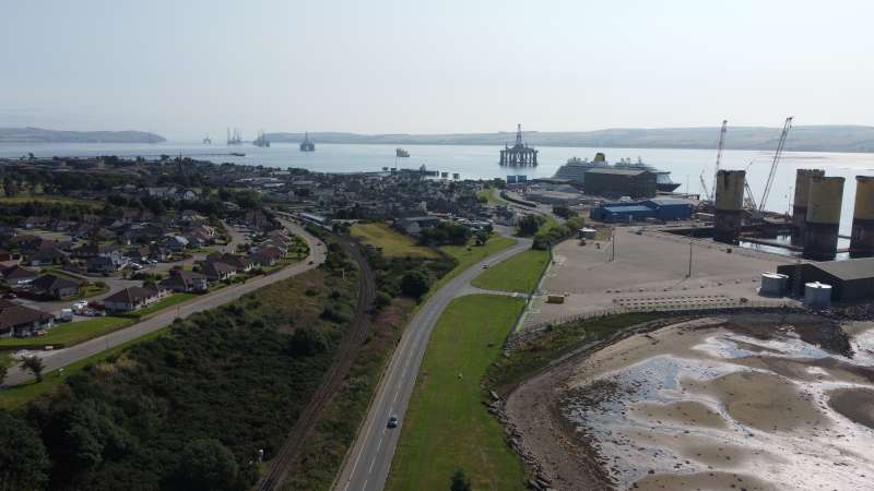 Aerial view of Invergordon on the Cromarty Firth, showing the town, coastal road and railway line alongside industrial port facilities. Offshore energy infrastructure and ships are visible in the harbour, with surrounding coastline.